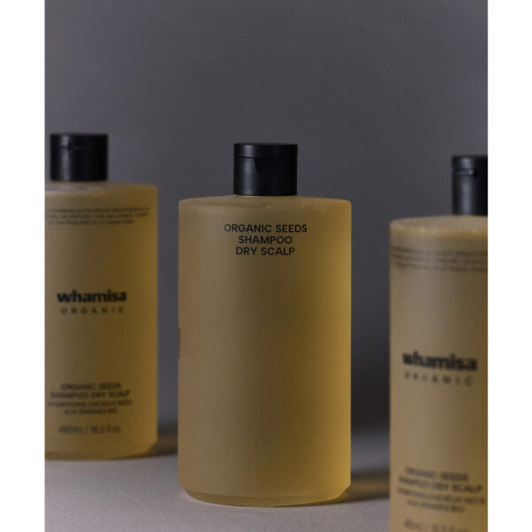 Whamisa Organic Seeds Shampoo Dry Scalp; kolme Whamisa Organic Seeds Shampoo -pulloa kuivalle päänahalle aseteltuna neutraalille taustalle; keskimmäinen pullo tarkasti fokuksessa, muut sumeina edessä ja takana; pullot mattapintaisia lieriömäisiä pyöristetyin olkaosin, tummanruskeita, valkoisin korkein; yksinkertaista valkoista groteskietikettitekstiä; ei ihmisiä tai käsiä kuvassa.