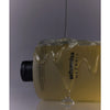 Whamisa Organic Seeds Shampoo Oily Scalp; Whamisa Organic -ihonhoitotuotteen pullo makaa kyljellään, hunajamainen neste valuu hellästi sen päälle ja kerääntyy alle; läpikuultava keltainen muovipullo lieriömäisenä, mustaa painettua etikettitekstiä yksinkertaisella groteskifontilla ja kiiltävä musta korkki; ei ihmisiä tai käsiä; tausta tasainen ja vaalea; kuva otettu hieman viistosti ylhäältä.