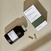 Wild Nutrition Ashwagandha Plus KSM-66 -ravintolisä; ruskea lieriömäinen pullo korkki irrallaan kahden beigen kapselin vieressä, lisäksi vihreä-valkoinen suorakaiteen muotoinen pahvirasia mattapinnalla, tekstissä KSM-66 Ashwagandha Plus selkeällä groteskifontilla; kaikki esineet sileällä beigellä alustalla, ei ihmisiä.