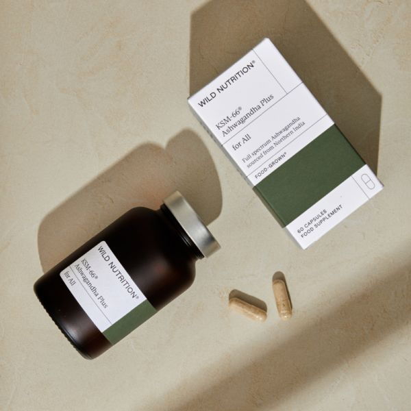 Wild Nutrition Ashwagandha Plus KSM-66 -ravintolisä; ruskea lieriömäinen pullo korkki irrallaan kahden beigen kapselin vieressä, lisäksi vihreä-valkoinen suorakaiteen muotoinen pahvirasia mattapinnalla, tekstissä KSM-66 Ashwagandha Plus selkeällä groteskifontilla; kaikki esineet sileällä beigellä alustalla, ei ihmisiä.