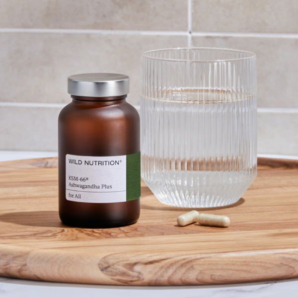 Wild Nutrition Ashwagandha Plus KSM-66 -ravintolisä; ruskea lieriömäinen lasipullo hopeisella kierrekorkilla, etiketissä mustaa groteskitekstiä Wild Nutrition KSM-66 Ashwagandha Plus; pullo suorakaiteen muotoisella puutarjottimella lasillisen vettä ja kahden beigen kapselin vieressä; vaalea, siisti tausta ilman ihmisiä.