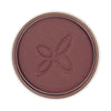 A round pan of mauve-colored powder blush features a subtle butterfly imprint in the center. Surface is matte with no visible shimmer. The product is inside a circular, metallic-looking compact without legible label text in frame. No hands or people are present. The background is plain and neutral, shot from above at close range.