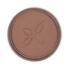 A round pan of brown pressed powder makeup with a subtle shimmer and a simple floral motif embossed in the center. The powder is contained in a circular metal pan, no lid or compact visible. There is no visible label text, typography, or brand marks. The surface shows a light-reflective finish. The background appears neutral and uncluttered with no people or hands present in the frame.