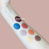An arm with eight eyeshadow swatches in colors: white, maroon (labeled BORDEAUX), blue, peach, silver, orange, gold, and brown is displayed against a pinkish background. Swatches are applied in parallel lines. One swatch has the word “BORDEAUX” printed in bold uppercase letters. The shot is close-up and does not show hands or packaging.