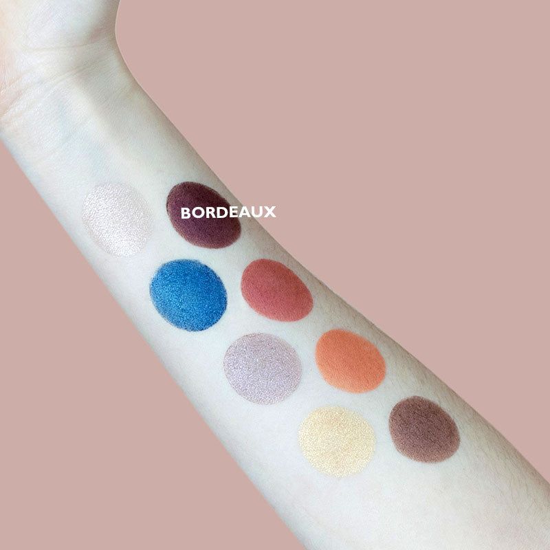 An arm with eight eyeshadow swatches in colors: white, maroon (labeled BORDEAUX), blue, peach, silver, orange, gold, and brown is displayed against a pinkish background. Swatches are applied in parallel lines. One swatch has the word “BORDEAUX” printed in bold uppercase letters. The shot is close-up and does not show hands or packaging.