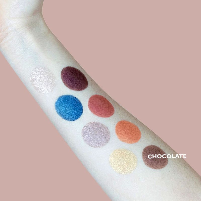An arm displays swatches of eight eyeshadow colors—white, burgundy, blue, pink, lilac, orange, gold, and brown—on a light skin tone. All swatches are applied in a row directly on the skin; finishes range from matte to shimmery. No packaging is visible. “CHOCOLATE” is printed near the brown swatch in uppercase block letters. Pale plain background; close-up shot.