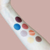 An arm displays eight eyeshadow swatches in various colors, including blue, brown, gold, peach, and plum, on a light skin tone. Swatches have both matte and shimmery finishes. The word SUNSET labels the peach shade in sans-serif capital letters printed directly above it. Eyeshadows form an even row from wrist to forearm. The background features a muted pink hue.