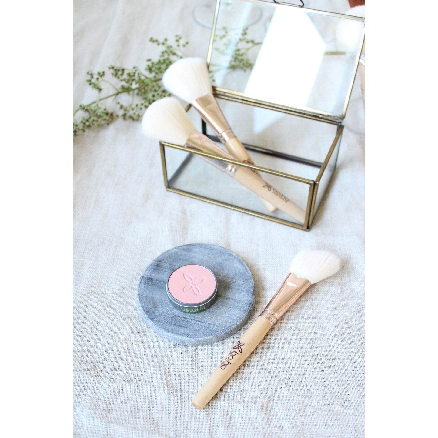 Boho Poskipuna Rose: meikkisivellin pellavaisella alustalla pyöreän puuterirasian vieressä, peilillinen suorakulmainen rasia sisältää kaksi lisää siveltimiä; heijastava metallireunainen kansi, sumea vihreys taustalla