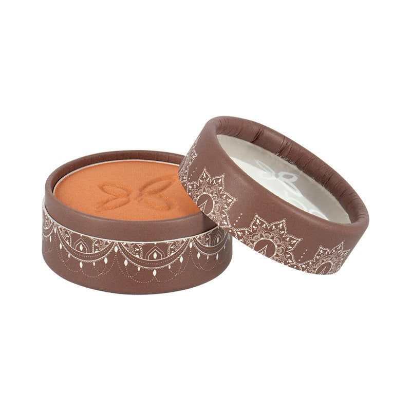 A round, brown cosmetic container with intricate white decorative patterns holds an orange-toned pressed powder. The lid, adorned with the same elegant design, is partially removed and rests beside the container. Both parts have a matte finish. There is no visible label or text on the packaging. The background is plain and untextured, with no people or hands present in the frame. This is a close-up product shot taken from above.
