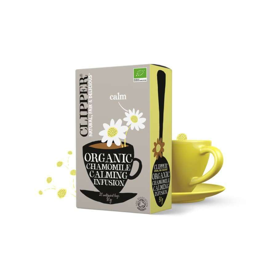 Clipper Organic Chamomile Calming Infusion -tee, päivänkakkarakuvituksin; rasia keltaisen teekupin ja tallrikan vieressä; suorakulmainen, pystyssä oleva rasia mattapinnalla, pääosin valkoinen ja keltainen; serif-fonttia pakkauksessa; teksti sisältää tuotenimen; neutraali tausta, ei ihmisiä kuvassa.