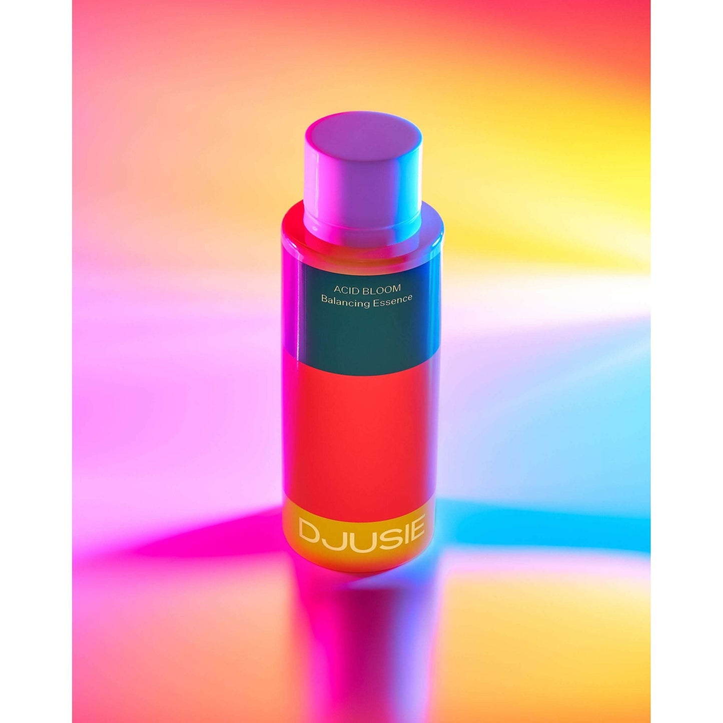 Djusie Acid Bloom Balancing Essence Hoitoneste; punainen ja pinkki pullo tekstillä "DJUSIE Acid Bloom Balancing Essence" alustalla, jota valaisee eloisa monivärinen valaistus keltaisen, pinkin ja sinisen sävyissä; pakkaus sylinterinmuotoinen kiiltäväpintainen lihavoiduin groteskitekstein; tausta tumma, heijastaen monivärisiä valoja alustalle pullon alla.