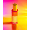 A bottle of DJUSIE Liquid Silk Perfect Cleansing Oil stands upright on a vibrant yellow and pink gradient background, casting a soft, luminous shadow. The bottle is cylindrical with a pump top and transparent body showing pale yellow liquid inside. The label displays product name in bold sans-serif text. No people or hands are visible in the image.