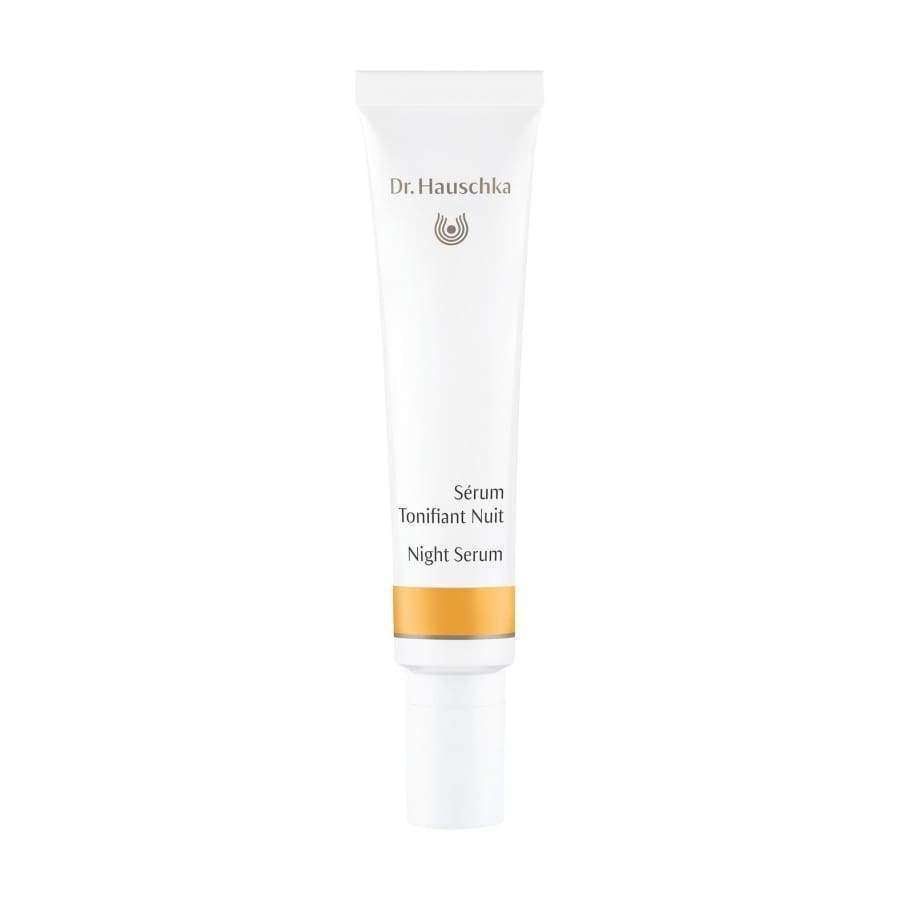 Dr. Hauschka Night Serum -tuubi, valkoinen tuubi kultaisin korostuksin ja kätevällä pumppuannostelijalla, pystyssä valkoista taustaa vasten; tuubi mattapintainen sylinterimäinen pyöristetyin reunoin, kultainen nauha annostelijan tyvellä; typografia siistiä ja minimalistista mustalla ja kultaisella; ei ihmisiä tai käsiä näkyvissä; kuva keskitetty ja hyvin valaistu täysin valkoista taustaa vasten.