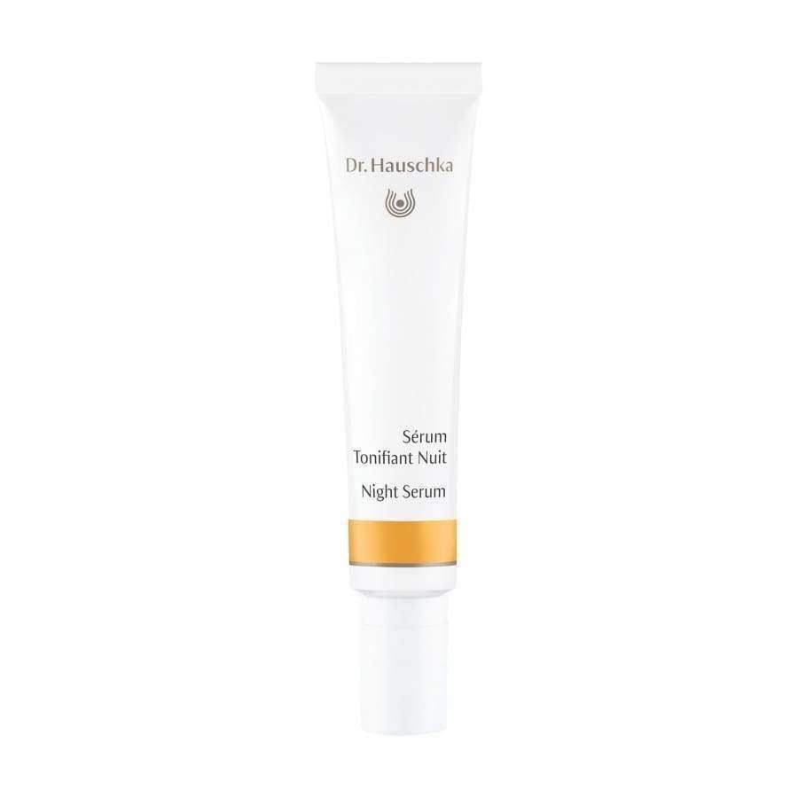Dr. Hauschka Night Serum -tuubi, valkoinen tuubi kultaisin korostuksin ja kätevällä pumppuannostelijalla, pystyssä valkoista taustaa vasten; tuubi mattapintainen sylinterimäinen pyöristetyin reunoin, kultainen nauha annostelijan tyvellä; typografia siistiä ja minimalistista mustalla ja kultaisella; ei ihmisiä tai käsiä näkyvissä; kuva keskitetty ja hyvin valaistu täysin valkoista taustaa vasten.