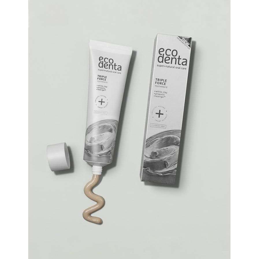 Ecodenta Triple Force Kolmivaikutteinen Hammastahna; tuubi Ecodenta Triple Force -hammastahnaa korkki irrotettuna, hieman luonnollista hammastahnaa puristettuna ulos, tuotepakkaus vieressä vaaleanharmaalla taustalla; tuubi lieriömäinen, mattapinta valkoisena ja vihreänä; korkki valkoista muovia; sekä tuubissa että rasiassa lihavoitua groteskitekstiä "Ecodenta Triple Force"; kuva otettu hyvin valaistuna ylhäältä, ei ihmisiä tai käsiä.