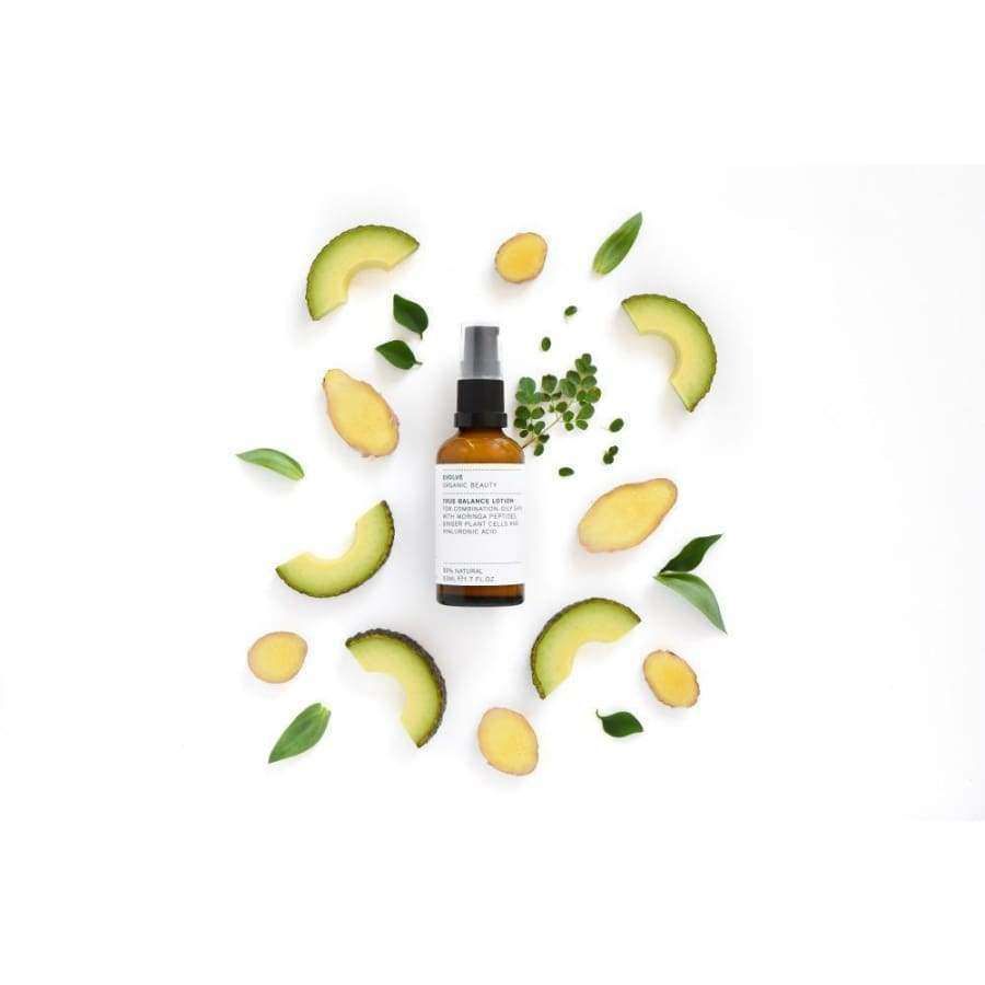 Evolve True Balance Tasapainottava Kasvovoide; ruskea suihkepullo muovisella liipaisimella, puolimatta pinta, ei näkyvää etikettiä tai brändimerkintää, lieriömäinen muoto; ympärillä siististi aseteltuja avokadoviipaleita, inkiväärinpaloja kuorineen ja hedelmälihoineen, litteitä vihreitä lehtiä ja pieniä vihreitä yrttioksia; kaikki aseteltu vaalealle taustalle ylhäältä kuvatussa otoksessa; ei ihmisiä tai käsiä.