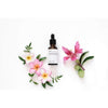 Evolve Nourishing Hair Elixir Kosteuttava Hiusseerumi -tippapullo valkoisella pinnalla, ympärillä värikkäitä vaaleanpunaisia ja valkoisia frangipanikukkia, tuuheita vihreitä lehtiä sekä hento vaaleanpunainen liljankukka; sylinterimäinen, kirkas ja kiiltäväpintainen pullo hopeanvärisellä korkilla ja lasisella tippaputkella; ei luettavaa etikettitekstiä tai brändäystä näkyvissä; ei ihmisiä tai käsiä kuvassa.