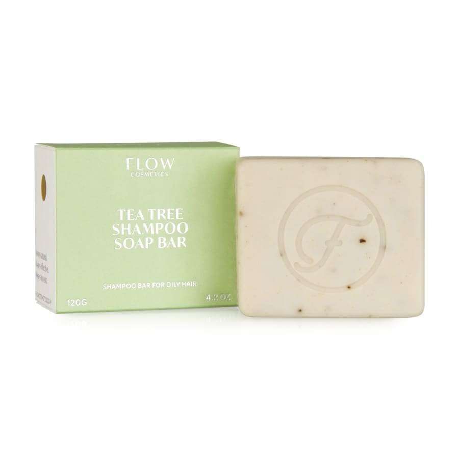Flow Cosmetics Tea Tree Shampoo Soap Bar; vaaleanvihreä rasia beigen saippuapalan vieressä, jossa kohokuvioitu pyöreä "F"-logo; pakkaus korostaa teepuuöljyn virkistäviä ominaisuuksia; rasiassa yksinkertaista mustaa groteskitekstiä kahdella painolla; molemmat mattapintaisia ja suorakulmaisia siistein reunoin; ei ihmisiä tai käsiä; tausta kauttaaltaan vaalea, ei lisärekvisiittaa.