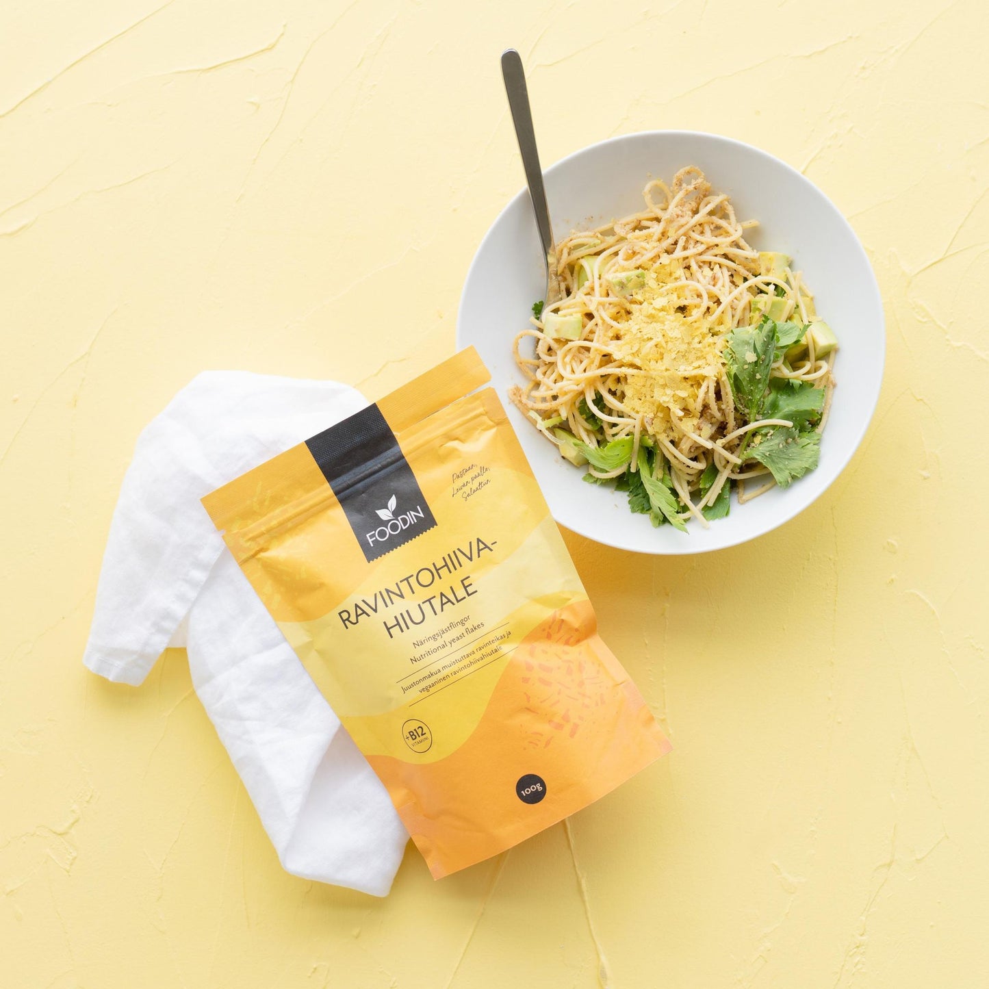 Foodin Ravintohiivahiutale -pussi; kulho pastaa vihreiden lehtien ja raastetun ravintohiivan kera keltaisella pinnalla, vieressä valkoinen lautasliina ja keltainen pussi tekstillä "Ravintohiiva Hiutale"; lusikka kulhossa; pussi mattapintainen, suorakulmainen, lihavoitua mustaa groteskitekstiä etusivulla; tasaisesti valaistu, kuvattu ylhäältäpäin, ei ihmisiä tai käsiä.