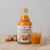 A bottle of turmeric juice with a label reading “Turmeric Juice” stands upright beside a clear glass containing the same bright yellow-orange liquid. Several whole, fresh turmeric roots are placed next to both. All items rest on a smooth, light-colored stone surface. No people or hands are visible in the frame. A bottle of turmeric juice labeled Turmeric Juice stands next to a glass filled with the vibrant drink, with fresh turmeric roots placed nearby on a light stone surface.