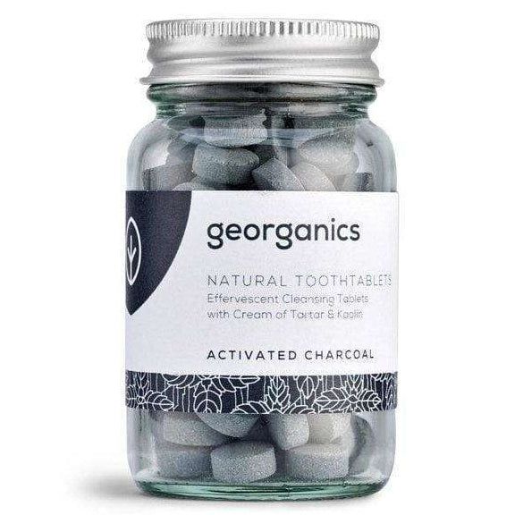 Georganics Hammastahnatabletit Aktiivihiili; lasipurkki tekstillä "Georganics Natural Tooth Tablets, Activated Charcoal", sisältää harmaita, pyöreitä tabletteja; purkki lieriömäinen mattaisella hopeanvärisellä metallikannella; etiketti valkoinen ja harmaa, groteskitypografiaa ja kasvikuvituksia alareunassa; tausta vaalea ja neutraali; ei ihmisiä tai käsiä kuvassa.