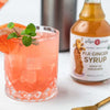 A pink grapefruit cocktail with a garnish of fresh mint and a grapefruit slice sits beside a bottle of Fiji Ginger Syrup on a white surface, showcasing vibrant flavors of citrus and ginger. The syrup is packaged in a clear glass bottle with a black cap and white label, featuring bold black serif typography reading “Fiji Ginger Syrup.” No people or hands appear.