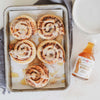 A tray of five iced cinnamon rolls on parchment paper, with ginger syrup drizzled on and around them. The rolls are round with white icing, arranged in a rectangular metal baking tray. A glass bottle labeled “Fiji Ginger Syrup” in a script font is placed upright nearby. Stacked glossy white ceramic plates are on a gray woven cloth background. No people or hands visible.