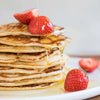 A stack of pancakes topped with fresh strawberries and syrup being poured over them, with extra strawberries scattered on the plate. Pancakes are round and golden brown with a matte finish; syrup is clear amber. No packaging or typography is visible. The background is a plain tabletop surface, shot from slightly above. No people or hands shown.