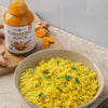 A bowl of vibrant yellow turmeric rice with green herb garnish is placed beside a clear glass bottle filled with golden-yellow liquid, labeled as turmeric juice in simple, sans serif font. Several slices of raw orange turmeric root are on the wooden board. The setting is a tabletop scene without people shown. A bowl of vibrant yellow turmeric rice garnished with herbs sits next to a bottle of golden turmeric juice and slices of fresh turmeric root on a wooden board.