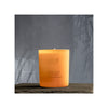 An orange cylindrical candle in a matte glass container shows the text “JOIK Home & Spa” and “Grapefruit & Mandarin” on a white rectangular label with simple sans-serif font. The candle sits on a smooth, dark surface. The background is gray with diffuse shadows of foliage. No people or hands are visible in the image.