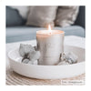 A lit JOIK Home & Spa candle in a cylindrical glass container with a matte white label displaying the product name in sans-serif type sits centered in a glossy white shallow dish. The dish is surrounded by decorative green leaves and a smooth, oval gray stone. The entire arrangement rests on a tan woven mat with visible texture. Background features an out-of-focus upholstered sofa in light color tones. No people or hands are present in the image.