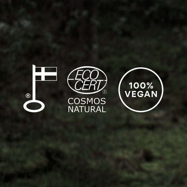 Taika Kosteuttava Käsivoide; kolme logoa tummalla taustalla: Pohjoismainen ympäristömerkki Joutsen, ECOCERT Cosmos Natural -sertifiointi ja "100% Vegan" -merkintä korostavat ekologisia ja luonnollisia tuotestandardeja; kaikki logot valkoisia selkein muodoin ja symbolein; tausta yhtenäisen musta, mattapinta; ei tuotteita, pakkauksia, ihmisiä tai esineitä kuvassa; lähikuva pelkistä logoista.