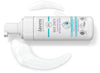 A plastic bottle of Lavera Basis Sensitiv Cleansing Foam with a white pump dispenser is positioned horizontally on a smooth, solid white surface. White foam is dispensed in curved formations near the bottle. The packaging is mainly white and blue with black sans-serif text and icons indicating organic, vegan certifications. No people or hands are visible in the image.