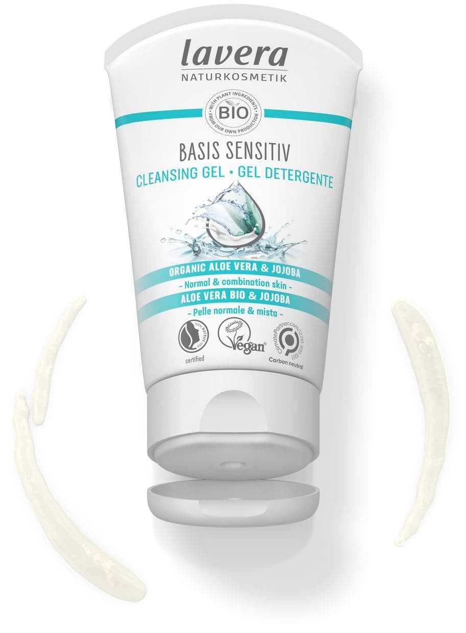 A tube of Lavera Basis Sensitiv Cleansing Gel with a splash of water, aloe, and jojoba illustrations is shown alongside product smears. The tube is white with blue-green accents and a glossy surface. Sans-serif black text on the label includes the product name and descriptors. The scene is well-lit with a plain background, flat lay composition, no people or hands present. Suitable for normal and combination skin, this vegan formula is also carbon neutral.