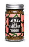 A glass jar of Littles Rich Hazelnut flavour infused instant coffee is shown. The jar has a reflective silver metal screw-on lid and a straight-sided cylindrical shape. Its front label is matte, with pink and green geometric patterns, and features legible product name text in white sans-serif font. The background is plain and well-lit; no people or hands are present in the image.