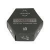 Kuusikulmainen musta laatikko merkinnällä Luonkos Complete Facial Oil Cleansing Cake for Men, jossa on pelkistettyjä viivapiirroksia kasvojenpuhdistusvaiheista sivuilla. Laatikko on mattamusta valkoisella pääteviivattomalla typografialla. Kuvitukset ovat yksinkertaisia valkoisia viivoja. Ihmisiä tai käsiä ei näy. Tuote on keskellä pelkistettyä, vaaleaa taustaa vasten tavanomaisessa tuotekuvassa.