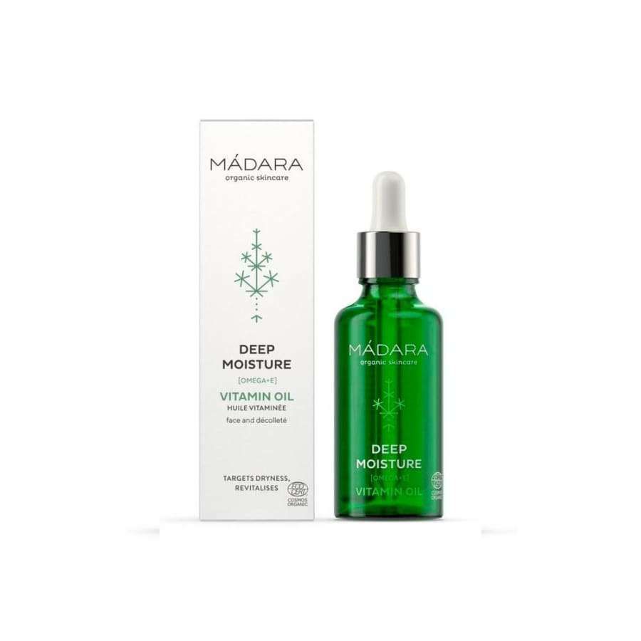 Madara Deep Moisture Vitamiiniöljy; vihreä lasinen tiputinpullo tekstillä "Mádara Deep Moisture Vitamin Oil" seisoo pakkauksensa vieressä, molemmat merkitty kasvojen ja dekolteen hoitoon; pakkaus minimalistinen mattapinnalla, lumihiutalekuvake ja yksinkertainen groteskityyppi; ei ihmisiä tai käsiä; tausta vaalea ja hyvin valaistu; kuva otettu edestä.