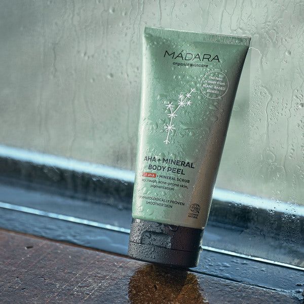Madara AHA + Mineraalikuorinta vartalolle; tuubi MÁDARA AHA+ Mineral Body Peel -tuotetta lepää märällä pinnalla ikkunan lähellä sadepisaroin; pakkaus vihreä mattapinnalla, sylinterinmuotoinen ja valkoinen groteskiteksti; kasviaiheisia kuvituksia painettu tuubiin; vain "MÁDARA AHA+ Mineral Body Peel" ja jotain pientä lisätekstiä on luettavissa etiketissä; ei ihmisiä tai käsiä kuvassa.