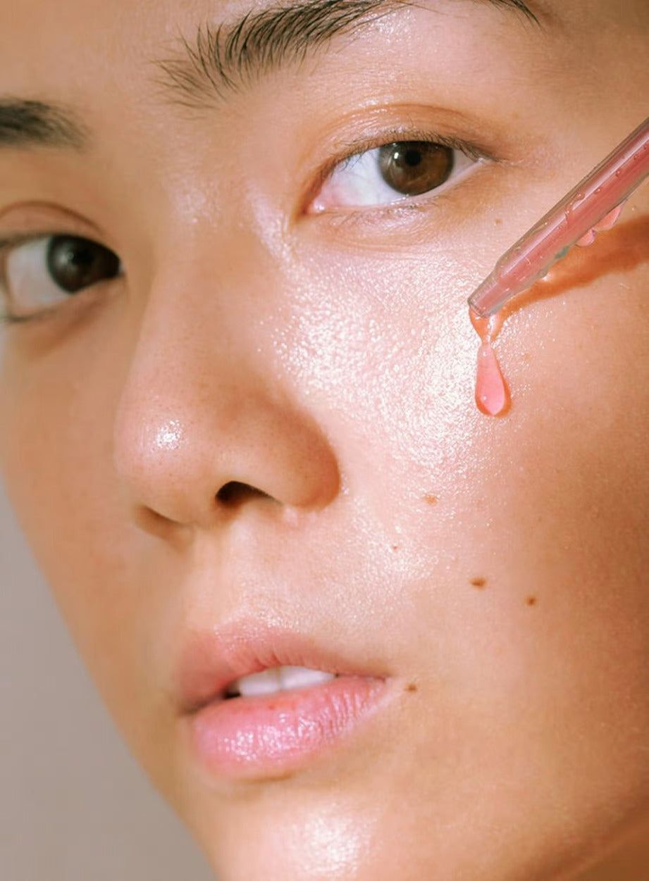 A close-up of a person’s face as a dropper applies pink serum to their cheek, highlighting smooth, hydrated skin and a natural glow. The dropper has a clear pipette with a white cap and contains translucent pink liquid. The background is solid white. No product packaging or label text is visible. Only the person’s cheek, lower face, and hand are shown in frame.