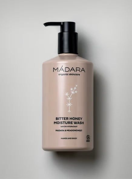 A beige bottle of Mádara Bitter Honey Moisture Wash with a black plastic pump is displayed on a smooth, light gray surface. The cylindrical bottle has a matte finish and features minimalistic white floral graphics. The label uses clean sans-serif typography, indicating it is for hands and body. No people or other objects are present in the shot.