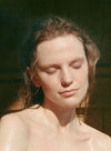 A woman with wet hair and closed eyes faces sunlight, her expression calm and serene. Her bare shoulders are visible. There is no visible product packaging, label, or surface material. The background is softly blurred in neutral tones. The shot is a close-up portrait focused on the subject’s face and upper shoulders.
