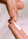 A close-up of a person applying a small amount of moisturizing cream onto the back of their hand using their fingers. The product is a white cream dispensed from an unseen container. The skin tone and texture on the hands are clearly visible. No packaging, labels, or branding are visible. The background is plain and out of focus.