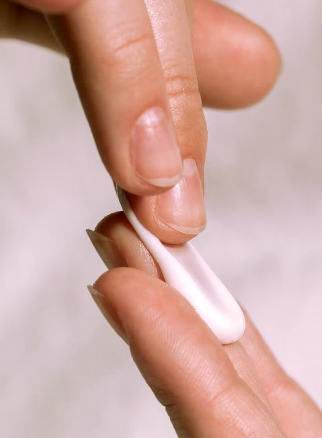A close-up of a person applying a small amount of moisturizing cream onto the back of their hand using their fingers. The product is a white cream dispensed from an unseen container. The skin tone and texture on the hands are clearly visible. No packaging, labels, or branding are visible. The background is plain and out of focus.