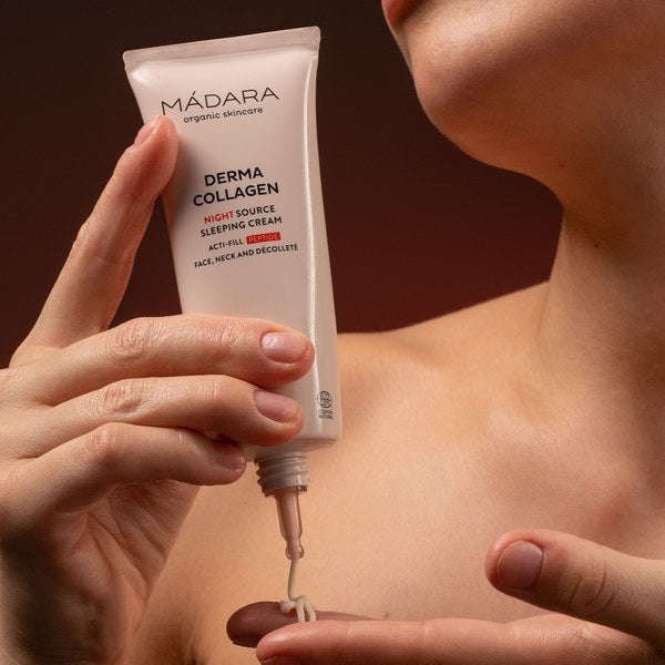 Madara Derma Collagen Night Source Yövoide; käsi puristaa tuubia tekstillä "MÁDARA Derma Collagen Night Source Sleeping Cream" sormenpäälle, kollageeni korostettuna ruskeaa taustaa vasten; tuubi matta ja sylinterinmuotoinen, valkoista groteskitekstiä eri kokoisin; pakkauksen väriä ei erikseen mainita; vain käsi ja sormenpää näkyvissä, ei muita esineitä tai ihmisiä; lähikuva.