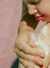 A close-up of a child washing their shoulder with soapy hands, bubbles covering the skin, and soft sunlight highlighting this gentle moment. No visible packaging, product branding, or label text. The scene is shot in natural lighting with a neutral background; only the child’s skin and hands are visible.