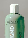 A close-up of a green MÁDARA organic skincare bottle with a white plastic cap. The bottle is cylindrical with a matte finish and displays the word “KIND” in bold uppercase letters along with eco-friendly icons printed on the label. Nourishing liquid is visibly dripping down the sides. There are no people, hands, or distractions in the frame; background is plain.