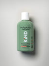 A green plastic bottle of MÁDARA organic skincare KIND Mild Shampoo for sensitive skin, suitable for kids and adults, is positioned horizontally on a light, solid-colored background. The cylindrical bottle has a matte finish with a flat cap. The label includes clear white sans-serif text and icons indicating formula features and skin safety. No people or hands are visible.