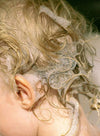 A close-up of the back of a child’s head with wet, curly blonde hair covered in soapy bubbles during a bath, capturing the playful moment. No visible product packaging or label text. Background is blurred and appears to be a bathroom setting, suggesting use of water and soap. The composition focuses on the child’s hair and soap bubbles.