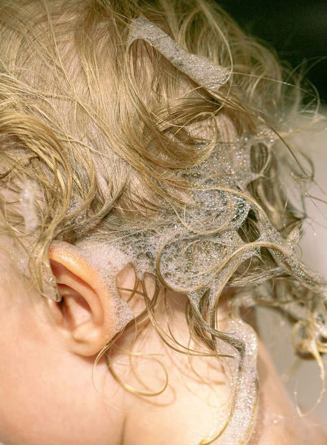A close-up of the back of a child’s head with wet, curly blonde hair covered in soapy bubbles during a bath, capturing the playful moment. No visible product packaging or label text. Background is blurred and appears to be a bathroom setting, suggesting use of water and soap. The composition focuses on the child’s hair and soap bubbles.