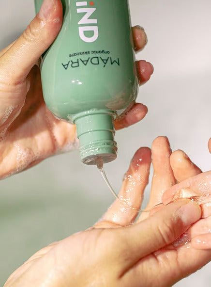 A person pours shampoo from a green MADARA bottle onto their hand, with a soft-focus background enhancing the soothing scene. The bottle is cylindrical with a matte green finish and features white uppercase sans-serif typography for the brand name “MADARA.” Only hands are visible, positioned in the foreground; no additional objects or label text are legible. The background is light-toned and out of focus.
